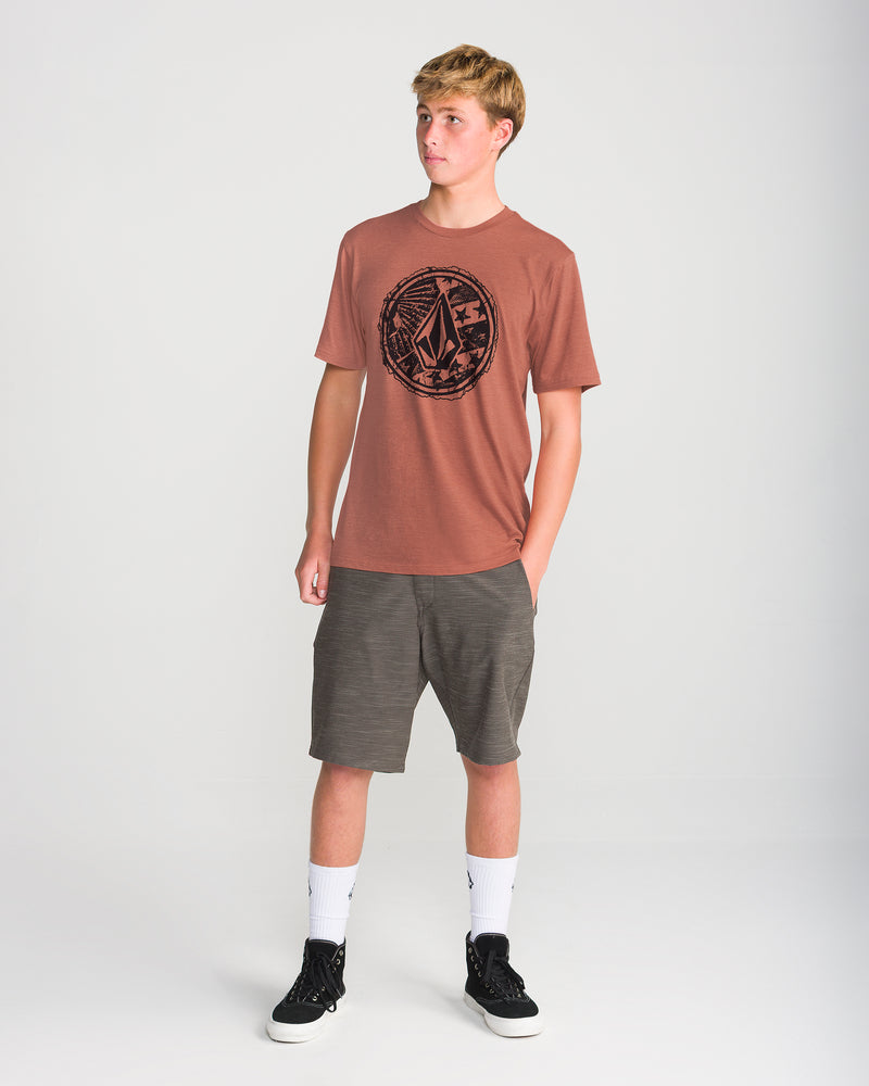 Teen boy stands against a white background, wearing the Volcom Men’s Core Stone Short Sleeve Tee in rust, gray shorts, white crew socks, and black sneakers, with one hand in his pocket looking left.