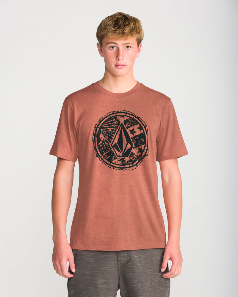 A young person wears the Volcom Men’s Core Stone Short Sleeve Tee in rust with a black geometric logo, paired with dark shorts, standing against a plain white background.
