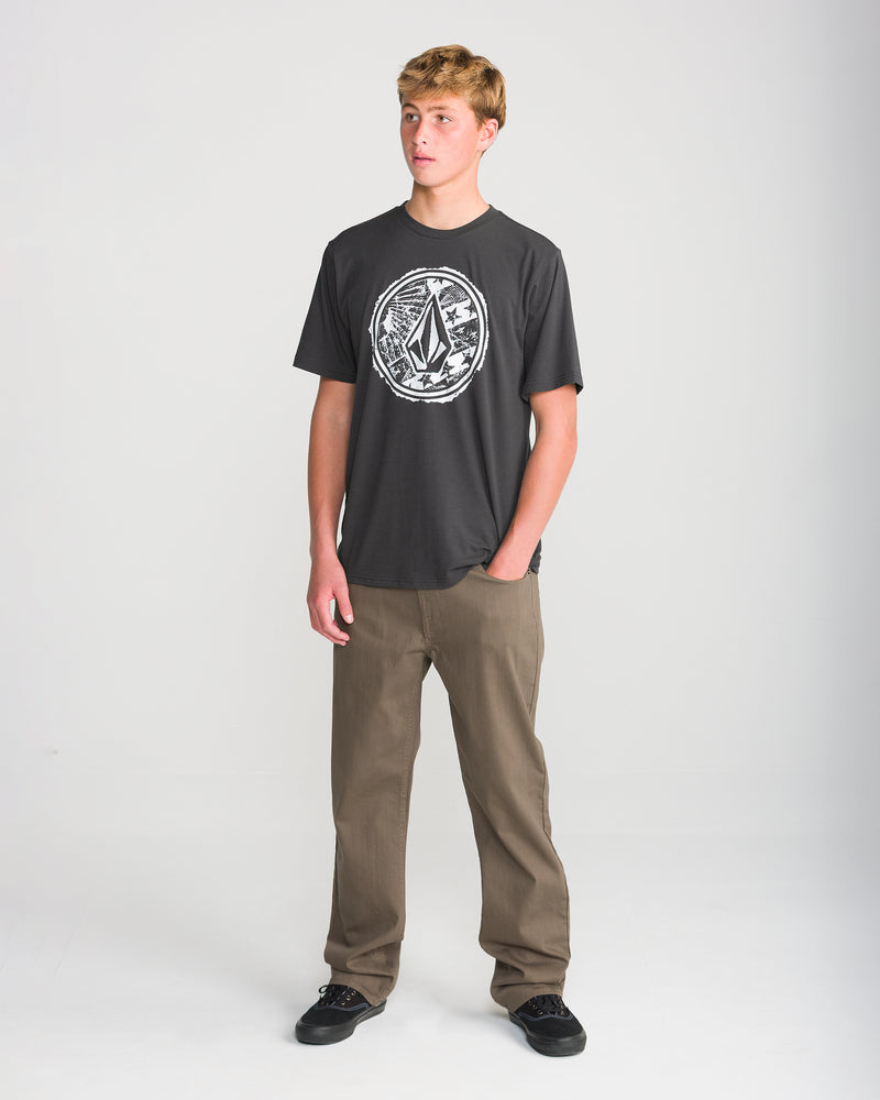 A young man stands against a plain background, wearing a Volcom Men’s Core Stone Short Sleeve Tee in Washed Black Heather, tan pants, and black shoes. He looks slightly left with a neutral expression and relaxed arms.