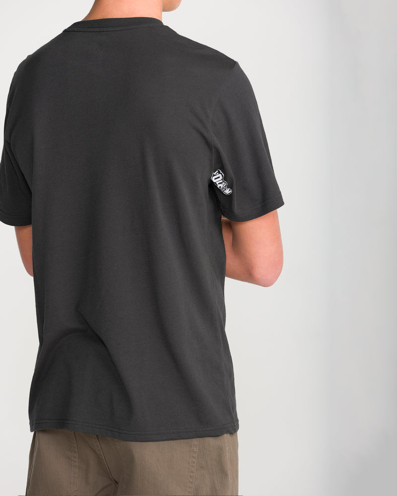 A person wears a Volcom Men’s Core Stone Short Sleeve Tee in Washed Black Heather with a modern fit and a small white tag on the sleeve, standing back to camera against a light gray background.
