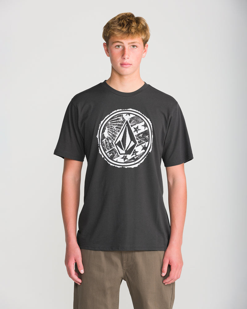Teen boy faces forward in a Volcom Men’s Core Stone Short Sleeve Tee, washed black heather with a large circular graphic, paired with brown pants against a plain light background for a modern fit look.
