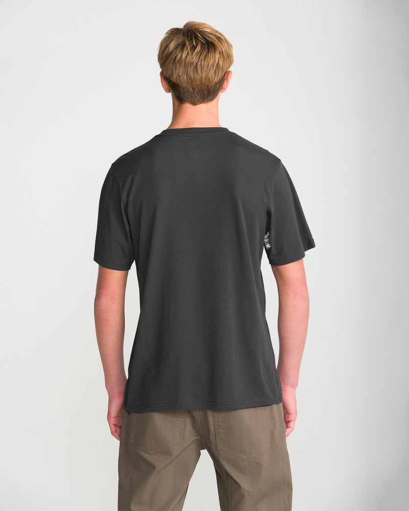 A person with short blond hair wears a Volcom Men’s Core Stone Short Sleeve Tee in washed black heather and khaki pants, standing with their back to the camera against a plain white background.