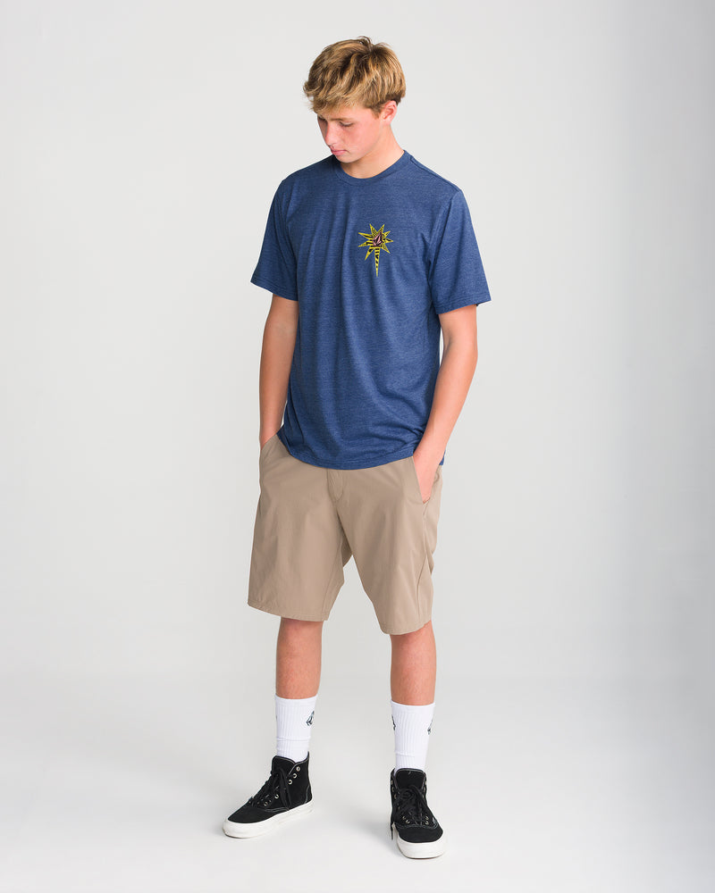 A teenage boy models the Volcom Men’s Op Blast Short Sleeve Tee in navy, paired with tan shorts, white socks, and black sneakers. He stands with hands in pockets, looking down against a plain light background.