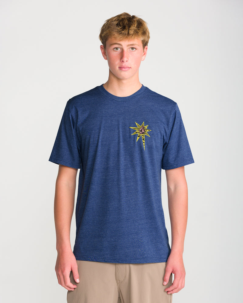 A young person faces forward in a Volcom Men’s Op Blast Short Sleeve Tee, a modern-fit navy shirt made from soft ring-spun cotton with a colorful starburst emblem, paired with light brown shorts against a plain background.