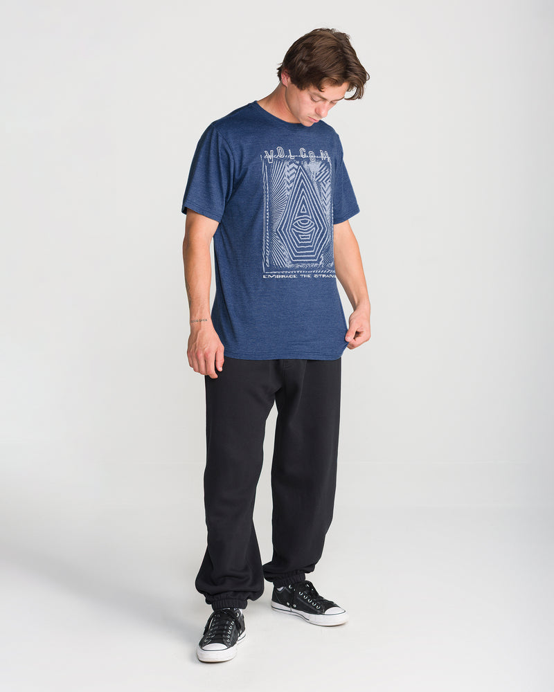 A person in the Volcom Men’s Insomniac Short Sleeve Tee, black pants, and black sneakers stands on a white background, looking down at their shirt.