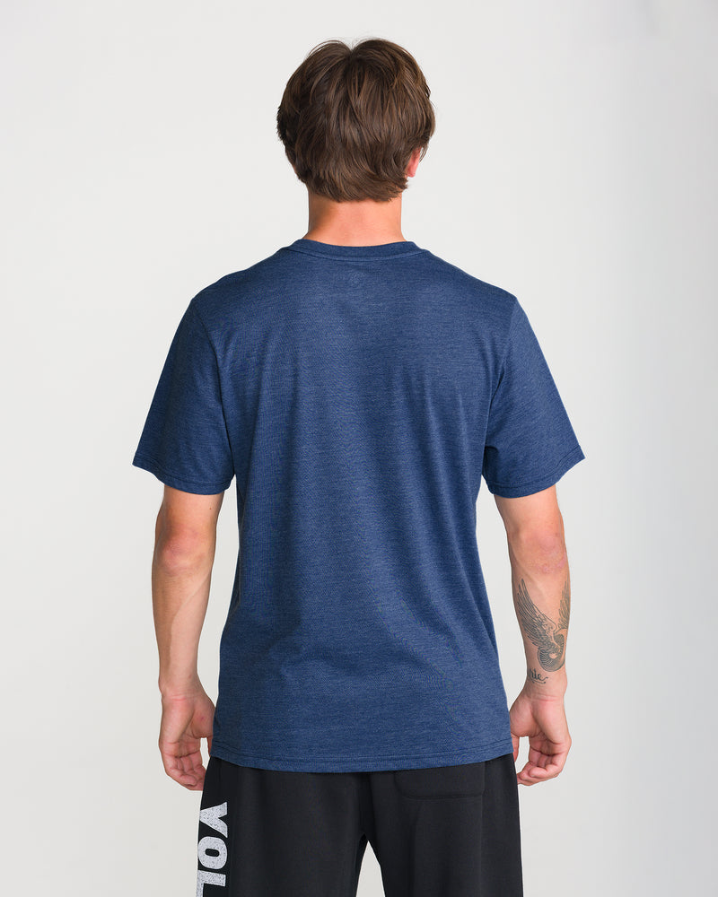 A person is shown from the back in a blue Men’s Insomniac Short Sleeve Tee by Volcom and black shorts, with a tattoo on their left forearm against a plain white background.