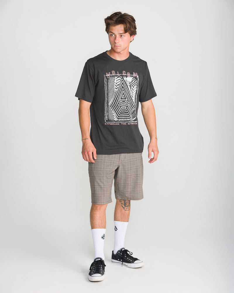 A young man wears the Volcom Men’s Insomniac Short Sleeve Tee in Washed Black Heather, plaid shorts, white logo socks, and black sneakers, gazing sideways with a neutral expression against a plain light background.