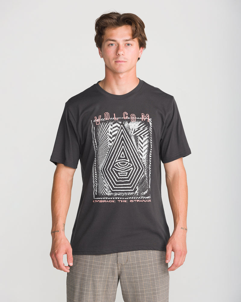 A person faces forward wearing the Volcom Men’s Insomniac Short Sleeve Tee in Washed Black Heather with geometric patterns and text, paired with plaid pants, against a plain light gray background.