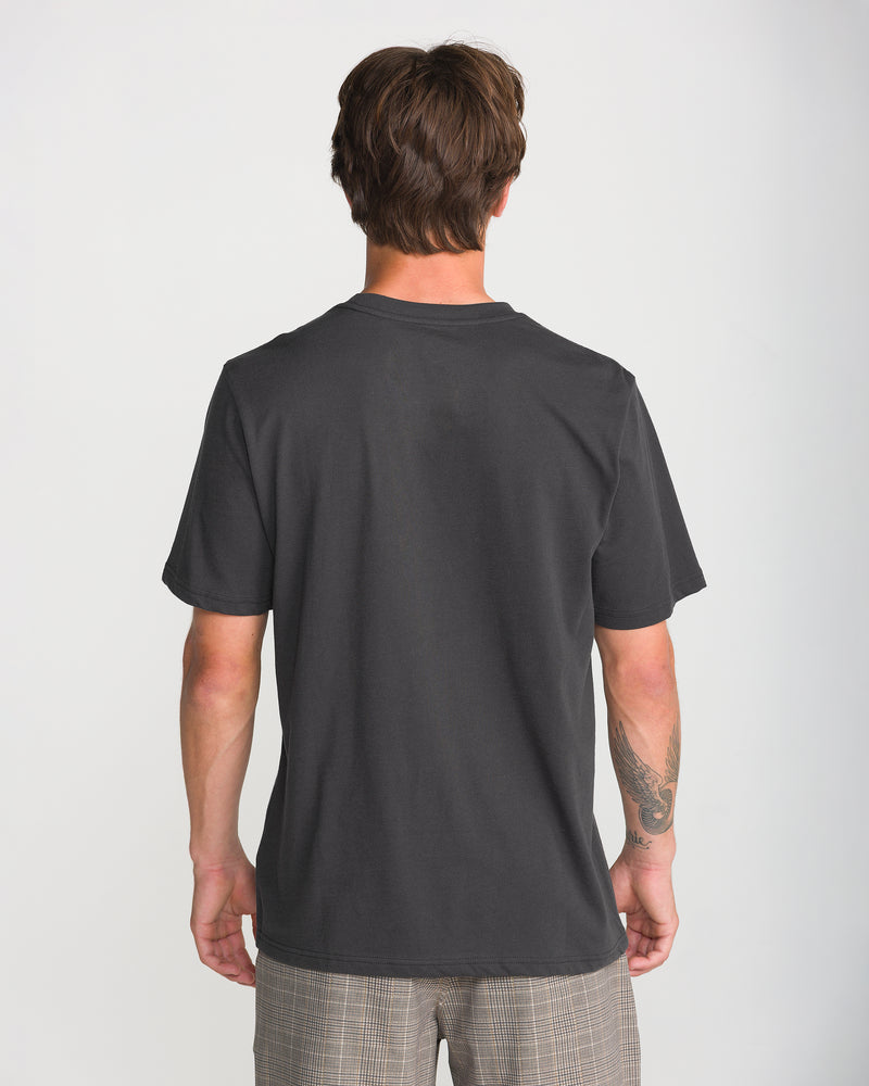 A person with short brown hair, wearing a Volcom Men’s Insomniac Short Sleeve Tee and plaid shorts, stands with their back to the camera against a light background.