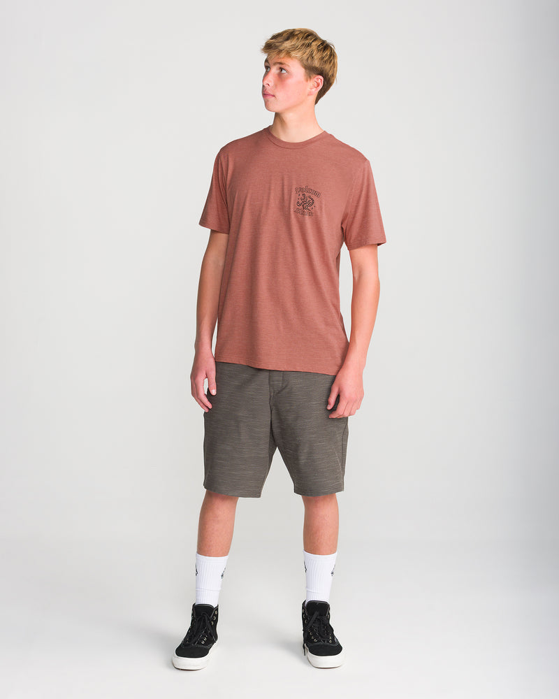 A young man wearing the Men’s Flash Short Sleeve Tee by Volcom in maroon, gray shorts, white crew socks, and black sneakers stands against a plain white backdrop, gazing to his left.