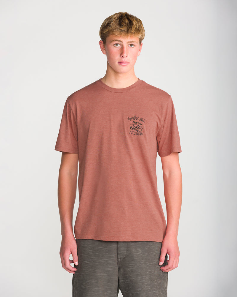 A young person with short blond hair wears the Volcom Men’s Flash Short Sleeve Tee in rust with a soft-hand screen print on the left chest and dark shorts, standing against a plain light background.