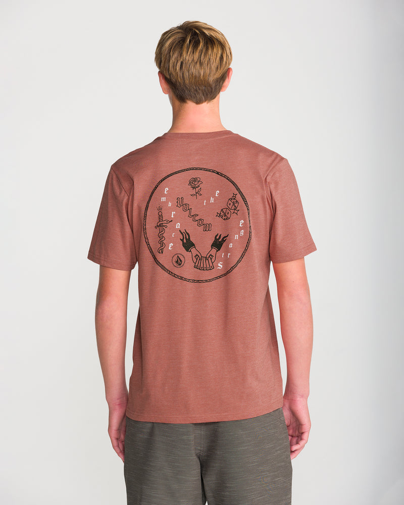 A person with short, light brown hair wears the Volcom Men’s Flash Short Sleeve Tee in reddish-brown with a bold abstract circular print on the back, standing against a plain white background.