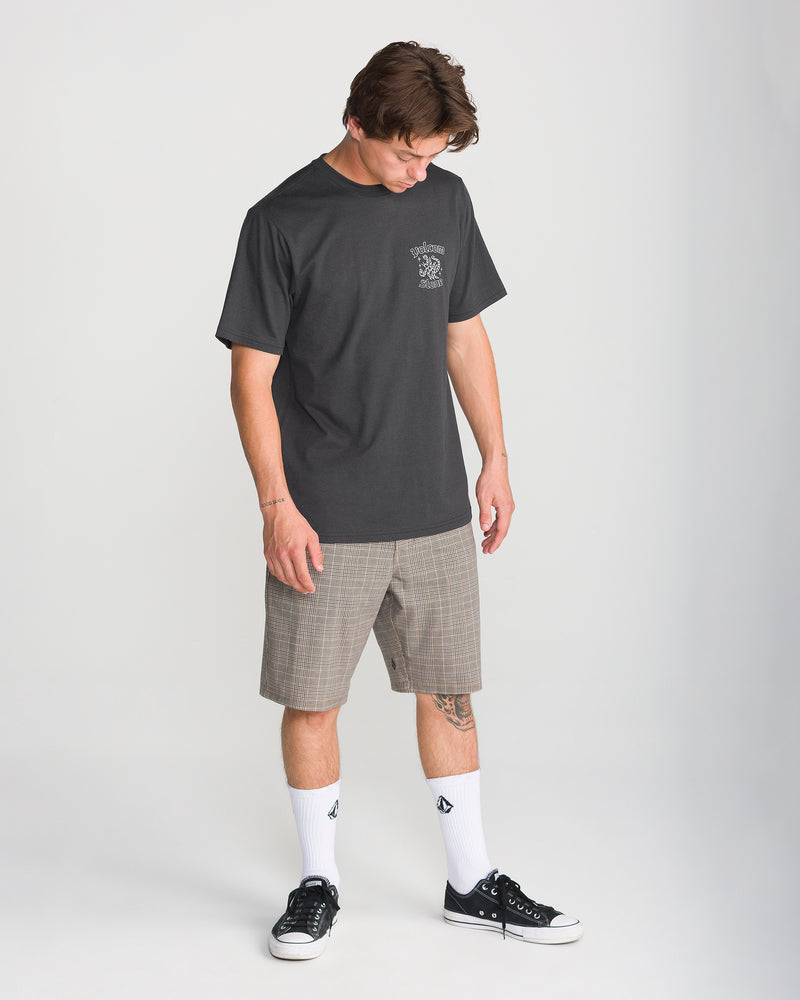 A person wearing a heathered dark grey Volcom Men’s Flash Short Sleeve Tee with a small chest graphic, tan plaid shorts, white socks, and black sneakers.