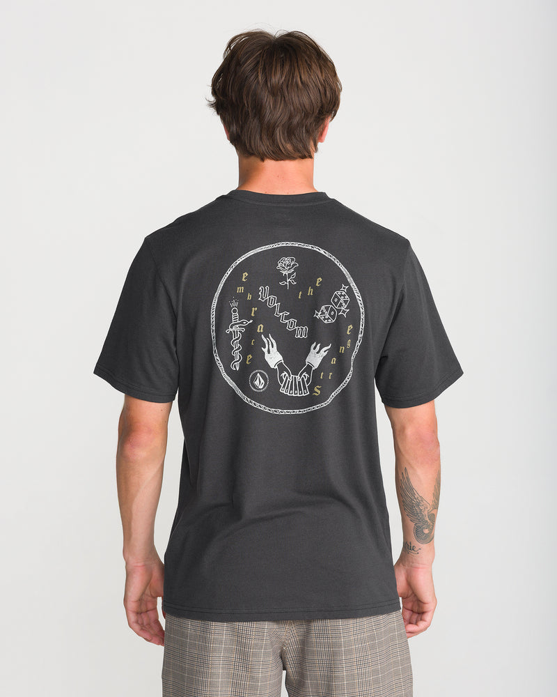 Back view of a person wearing a heathered grey Volcom Men’s Flash Short Sleeve Tee with a white circular graphic on the back featuring a dagger, dice, flames, a rose, and circular text.