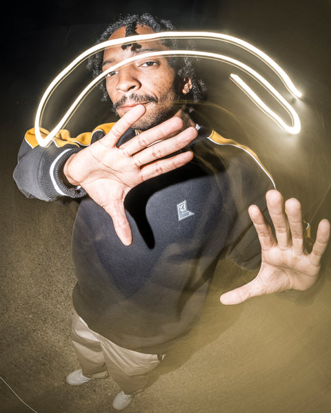 A person stands with hands raised toward the camera at night, a light trail creating an arc above their head. They are wearing the Volcom Men’s Flying Hi Crew, a hoodie with colorblocked sleeves crafted from soft REPREVE recycled polyester brushed-back fleece, and are captured in an image featuring a distorted, wide-angle effect.