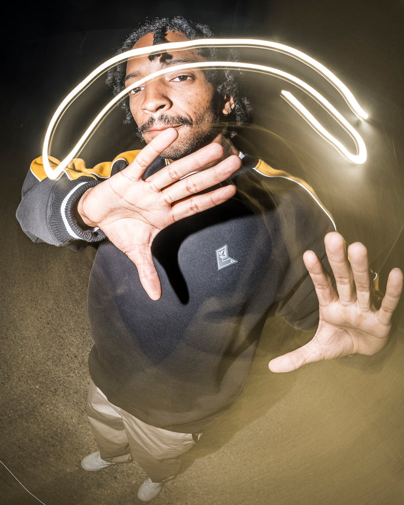 A person stands with hands raised toward the camera at night, a light trail creating an arc above their head. They are wearing the Volcom Men’s Flying Hi Crew, a hoodie with colorblocked sleeves crafted from soft REPREVE recycled polyester brushed-back fleece, and are captured in an image featuring a distorted, wide-angle effect.