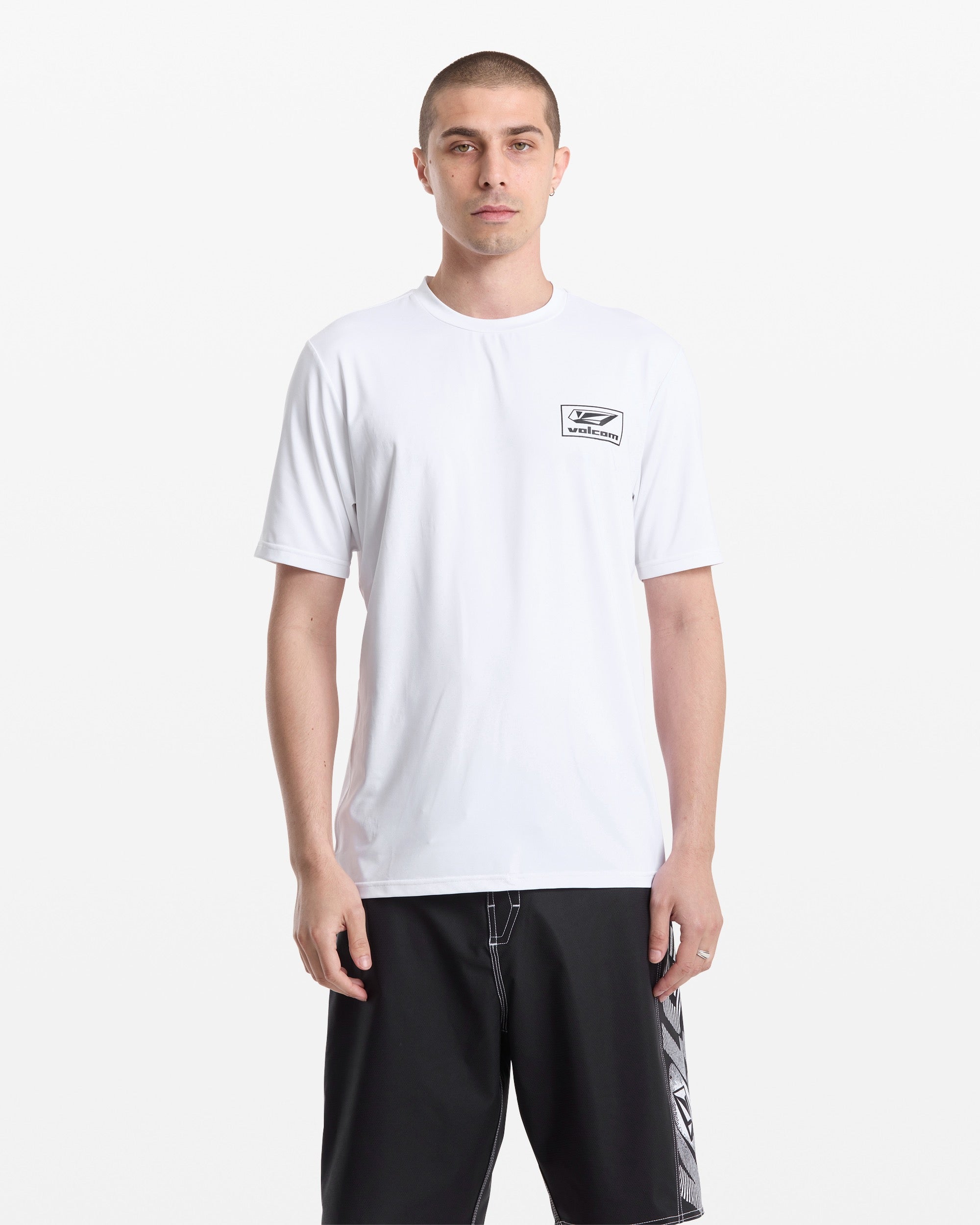 A person with short hair wears a plain white Volcom Men's Modstone Loose Short Sleeve Surf Tee and black shorts with a patterned panel, standing against a white background.