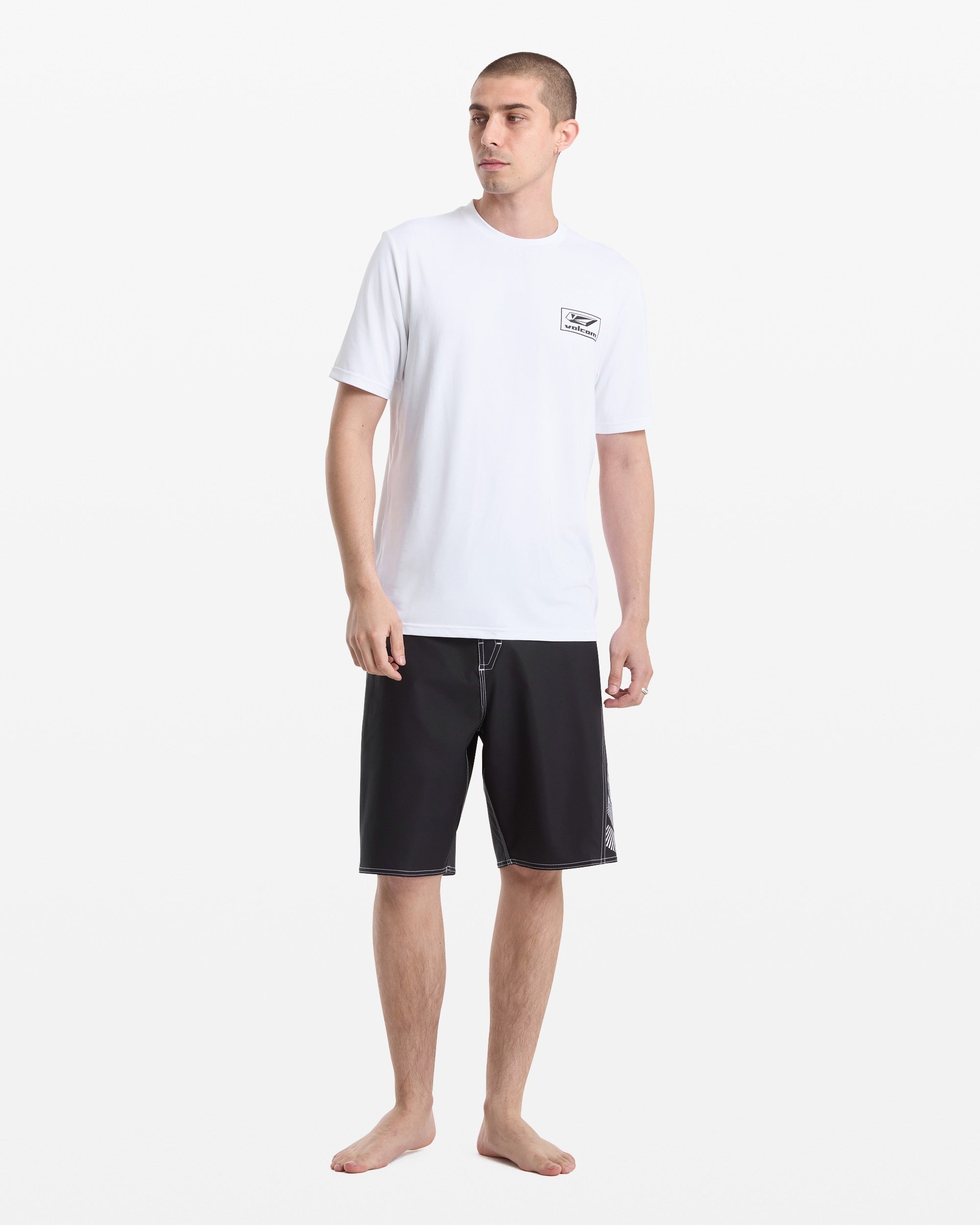 A man stands barefoot against a white background, wearing the Volcom Men's Modstone Loose Short Sleeve Surf Tee with UV protection and chest logo, paired with black knee-length shorts featuring white stitching.