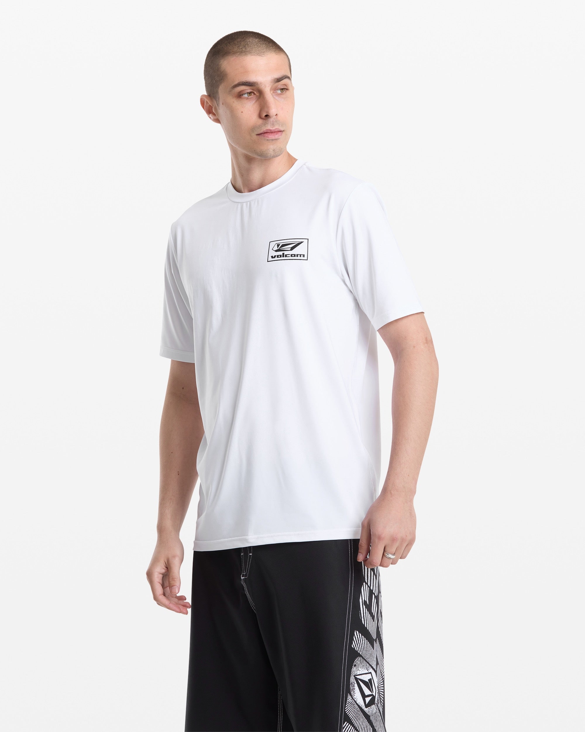 A person with short hair wears the Volcom Men's Modstone Loose Short Sleeve Surf Tee, a relaxed fit white T-shirt with a small chest graphic, paired with bold-patterned black pants, standing against a plain white background.