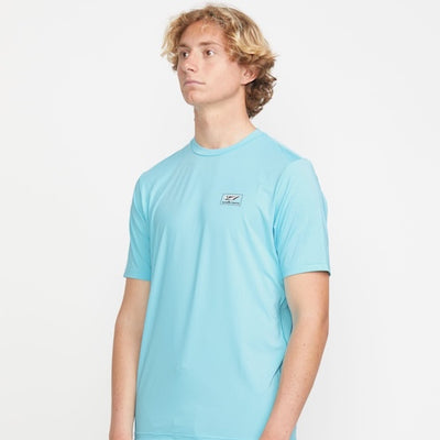 A young man with wavy blond hair wears a light blue short-sleeve t-shirt with a small rectangular logo on the chest, standing against a plain white background.