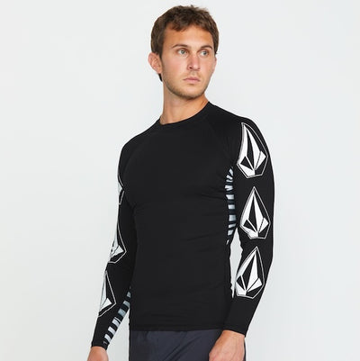 A man wearing a fitted black long-sleeve shirt with white geometric logo designs on the sleeves stands facing slightly to the side against a plain white background.