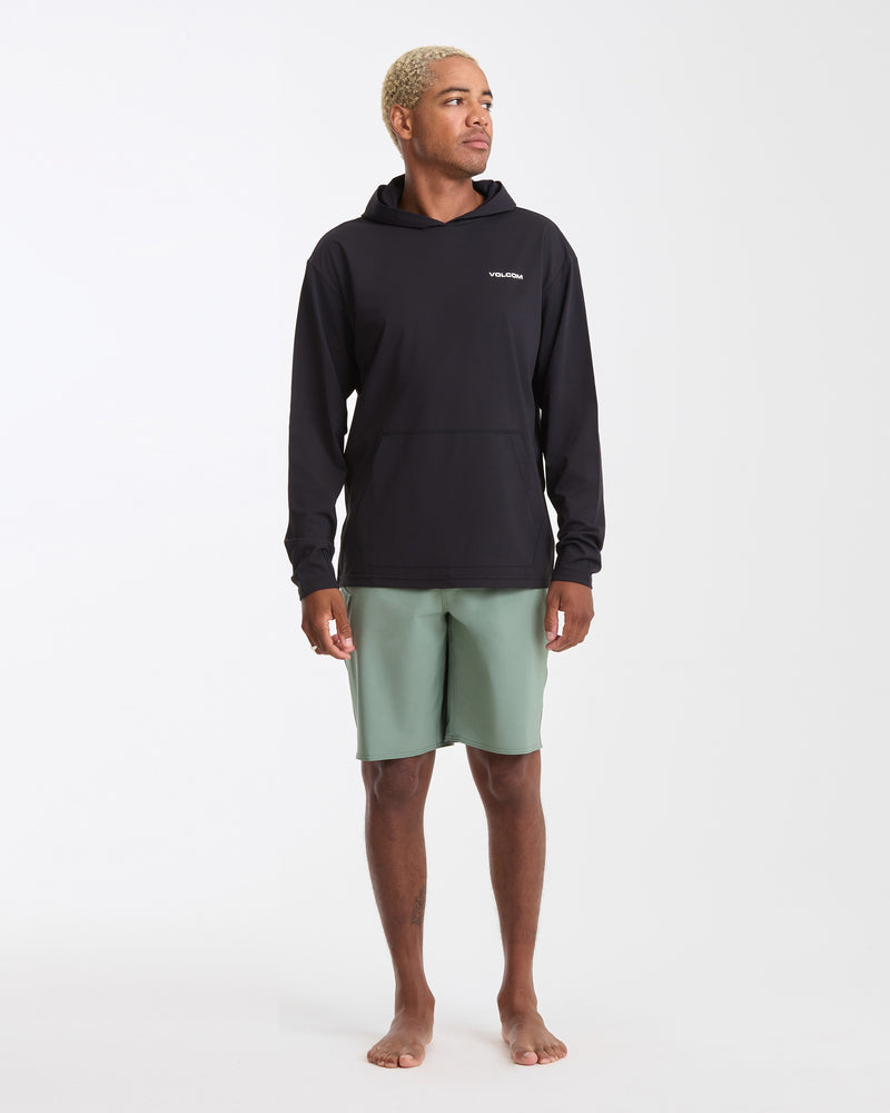 A person with short blond hair stands barefoot on a white background, wearing a Volcom Men's Pullover Hood Long Sleeve in black with UV protection and light green shorts, looking slightly to the side.