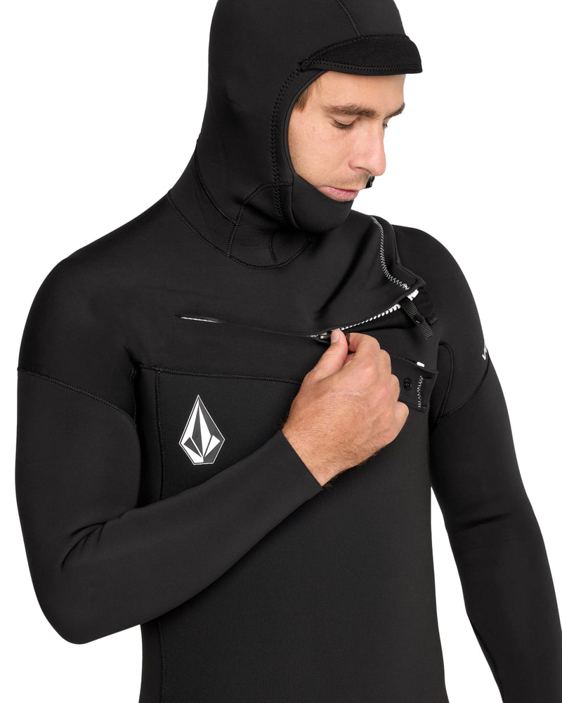 A person is zipping up the chest of a Volcom Men's 4/3MM Hooded Chest Zip Full Wetsuit made from limestone-based neoprene, featuring an attached hood and a white geometric Volcom logo, against a plain white background.