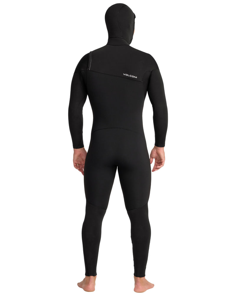 A barefoot person faces away, wearing the Volcom Men's 4/3MM Hooded Chest Zip Full Wetsuit. This full-body black wetsuit has long sleeves, a thermal rapid-dry lining, and VOLCOM branding on the upper back.