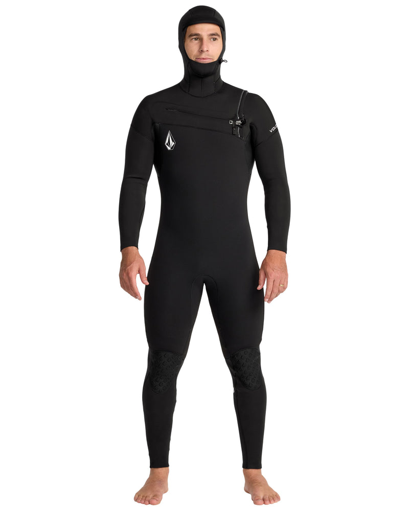 A person stands barefoot in the Volcom Men's 4/3MM Hooded Chest Zip Full Wetsuit—made with limestone-based neoprene. The black full-body suit features subtle chest and arm branding, reinforced knees, and an attached hood.