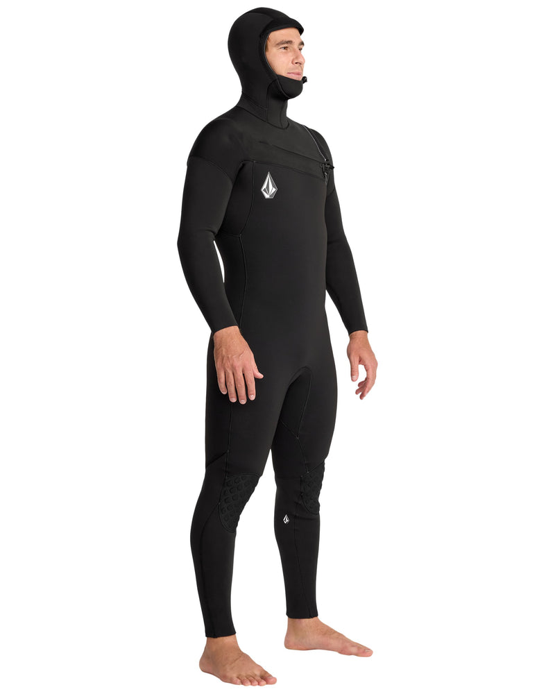 A person stands barefoot in the Volcom Men's 4/3MM Hooded Chest Zip Full Wetsuit—black, full-body, limestone-based neoprene with a chest logo, hood, and rapid-dry thermal lining—facing slightly right. Suitable for cold water.