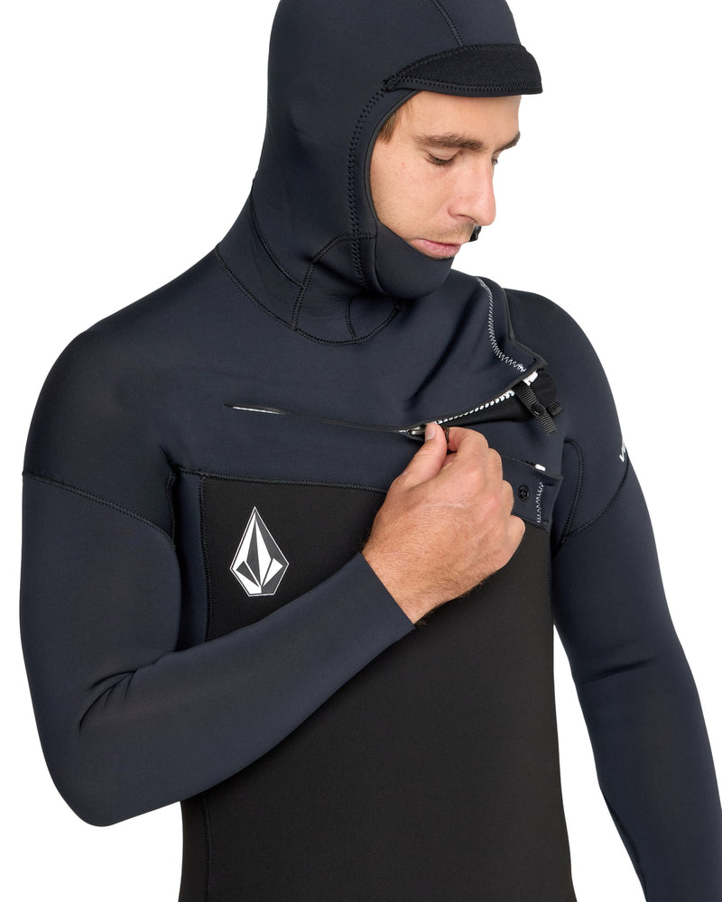 A person wearing the Volcom Men's 5/4/3 Modulator Hooded Chest Zip Full Wetsuit made from limestone-based neoprene unzips the front, revealing a white geometric logo on the left chest while looking down at the zipper.