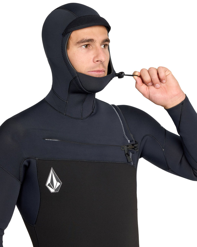 A person in the Volcom Men's 5/4/3 Modulator Hooded Chest Zip Full Wetsuit, made from limestone-based neoprene and featuring a geometric chest logo, pulls a drawstring near their chin.