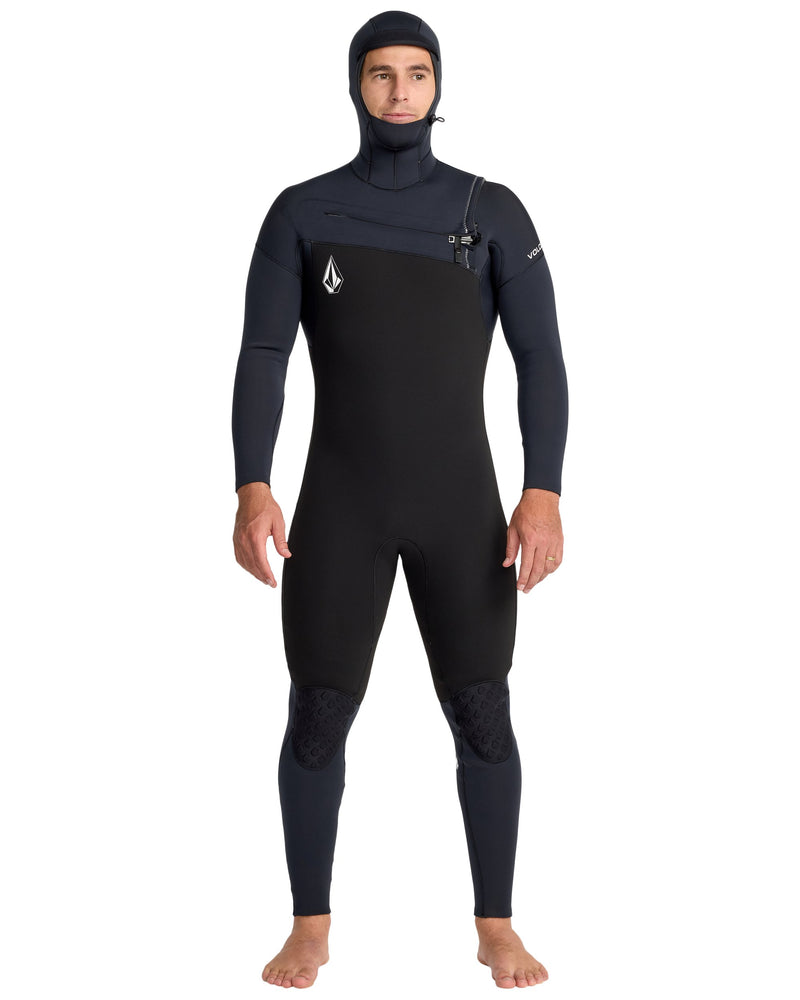 A man stands barefoot facing forward, arms relaxed, wearing the Volcom Men's 5/4/3 Modulator Hooded Chest Zip Full Wetsuit in black. Made from limestone-based neoprene, it has a chest logo and reinforced knee panels.