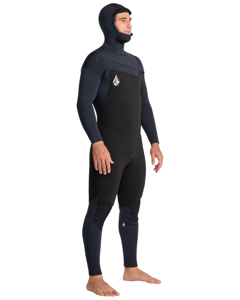 A man stands barefoot on a white background, wearing the Volcom Men's 5/4/3 Modulator Hooded Chest Zip Full Wetsuit in black, made from limestone-based neoprene, looking slightly to the side.