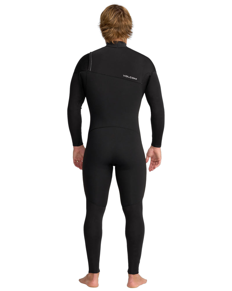 A person with short light brown hair stands barefoot, facing away, wearing the Volcom Men's 3/2 Modulator Chest Zip Full Wetsuit with VOLCOM on the upper back.
