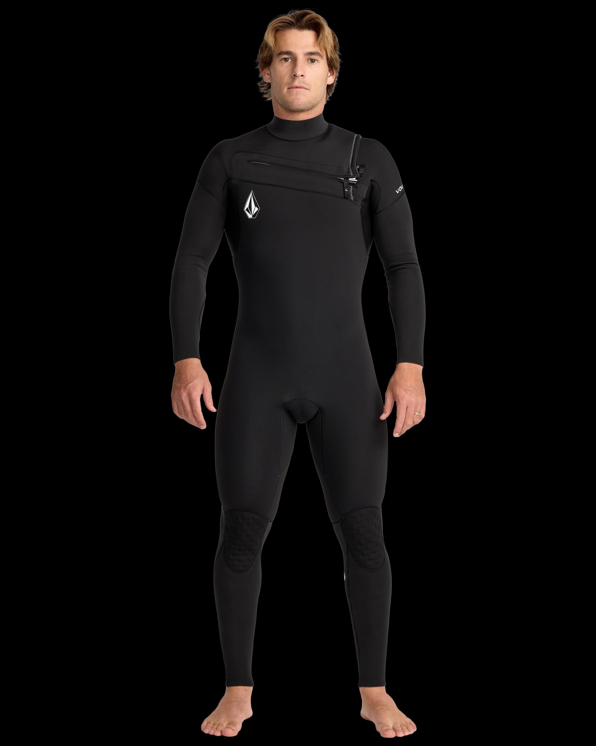 A man stands barefoot against a black background, wearing the Volcom Men's 3/2 Modulator Chest Zip Full Wetsuit, featuring a front zip and white Volcom logos on the chest and shoulder.