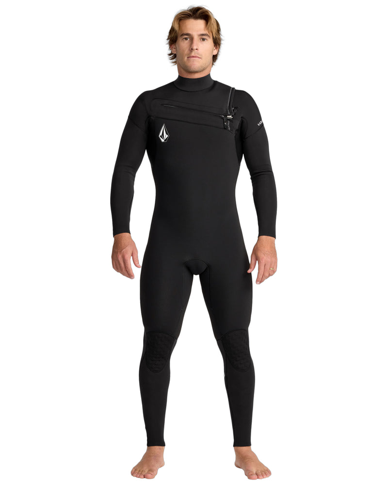 A man stands barefoot facing forward in the Volcom Men's 3/2 Modulator Chest Zip Full Wetsuit, featuring chest zipper and logo details on the chest and sleeve, against a plain white background.