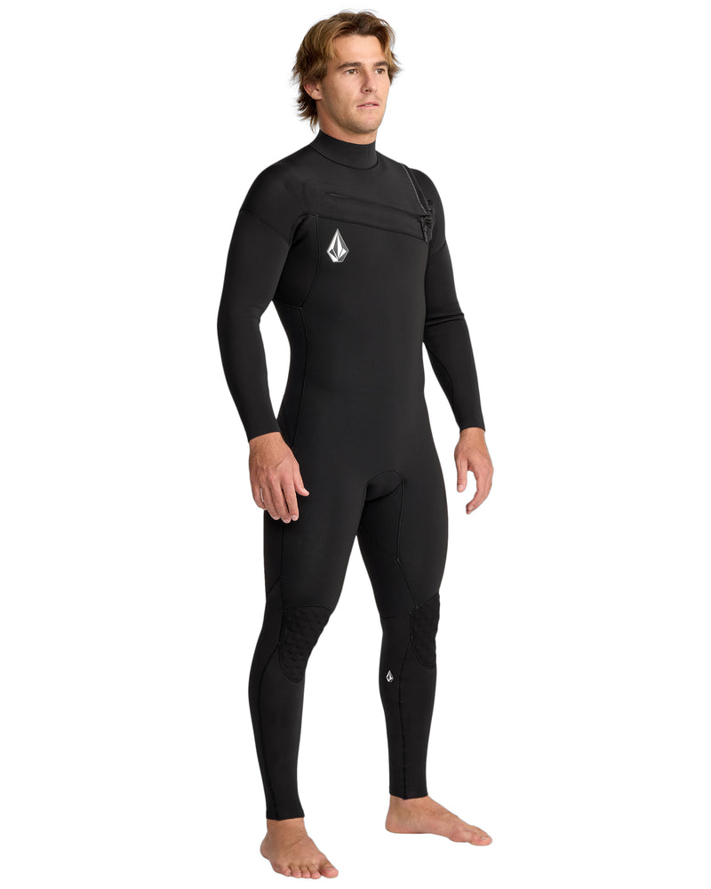 A man stands barefoot in a Volcom Men's 3/2 Modulator Chest Zip Full Wetsuit, featuring long sleeves, a chest zipper, and small Volcom logos on the chest and knee. The background is plain white.