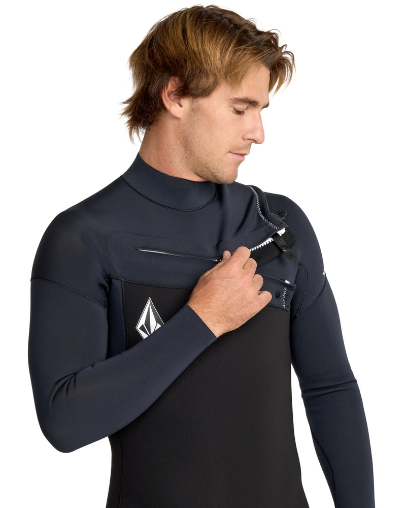 A man with light brown hair, wearing the Volcom Men's 4/3 Modulator Chest Zip Full Wetsuit, pulls down the chest zipper while looking at it. A white logo is visible on his chest. The background is plain white.