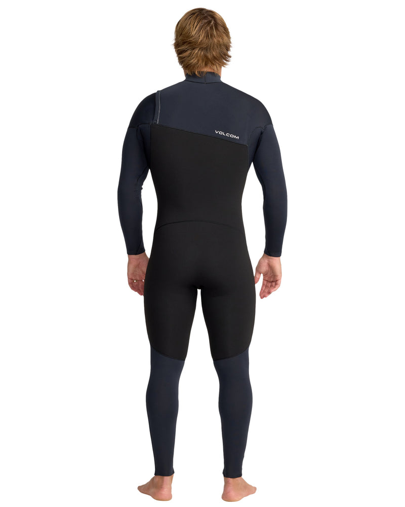 A person with light brown hair, barefoot and facing away, wears the Volcom Men's 4/3 Modulator Chest Zip Full Wetsuit in black and dark navy, featuring long sleeves and the Volcom logo on the upper back.