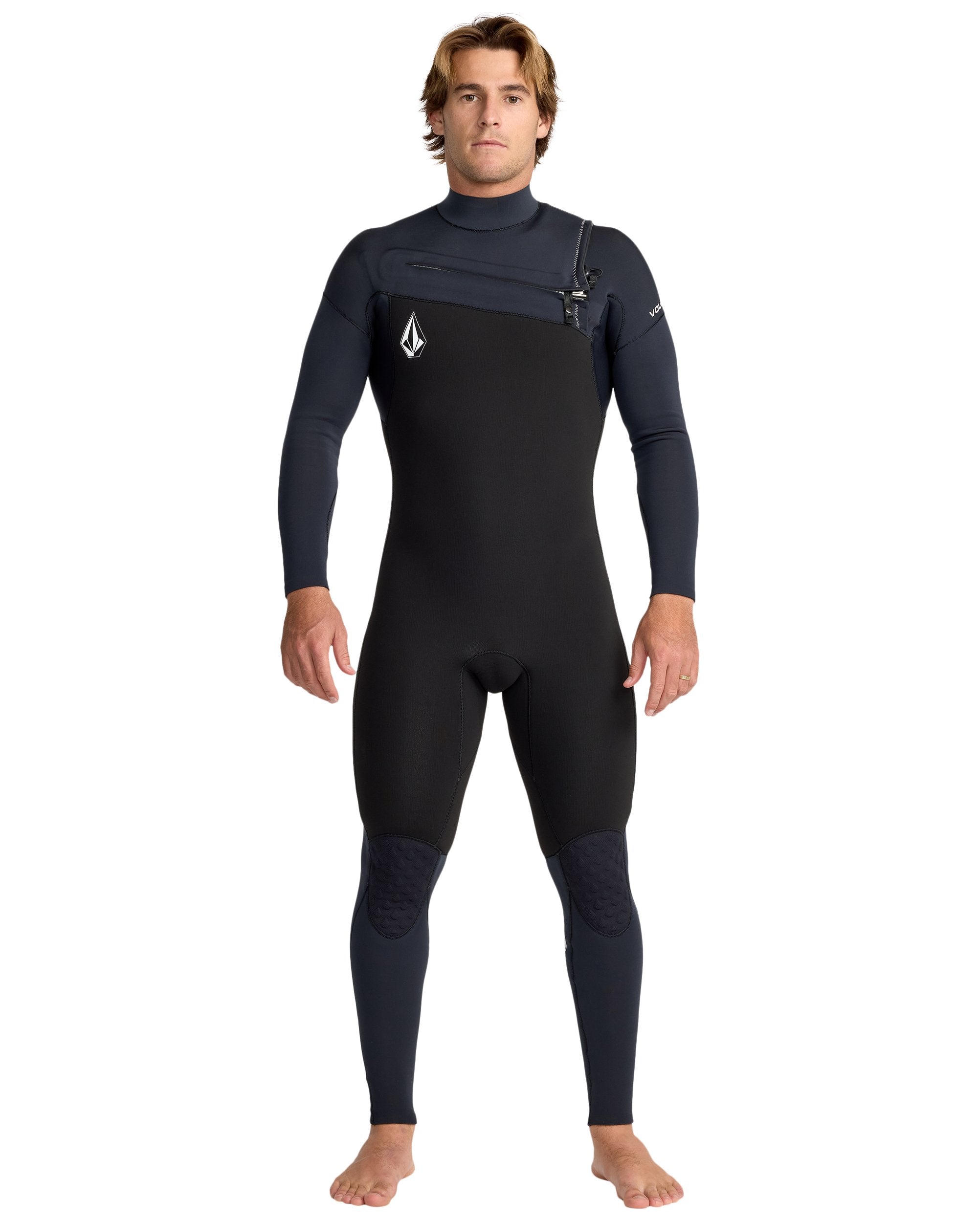A man with light brown hair stands barefoot, facing forward, in the Volcom Men's 4/3 Modulator Chest Zip Full Wetsuit—black and dark gray with long sleeves and a small logo on the chest. His arms are relaxed at his sides.
