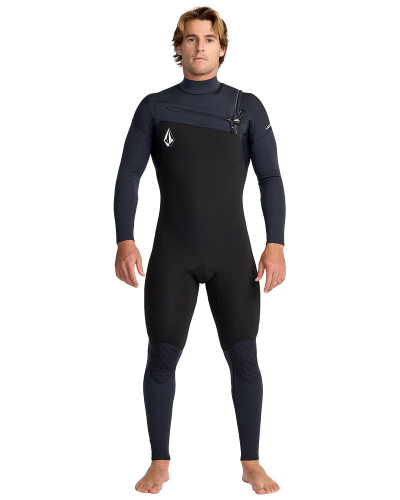A man with light brown hair stands barefoot, facing forward, in the Volcom Men's 4/3 Modulator Chest Zip Full Wetsuit—black and dark gray with long sleeves and a small logo on the chest. His arms are relaxed at his sides.
