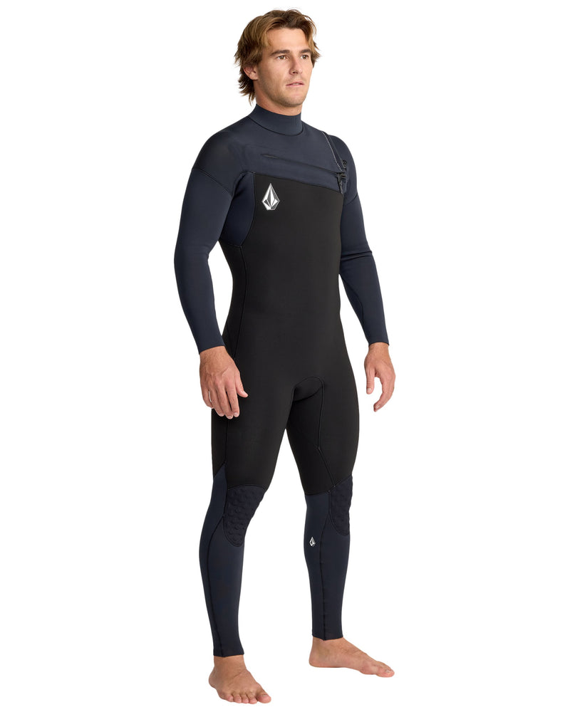 A man stands barefoot, slightly turned, against a white background wearing the Volcom Men's 4/3 Modulator Chest Zip Full Wetsuit in black and dark blue, featuring long sleeves and chest and knee logos.