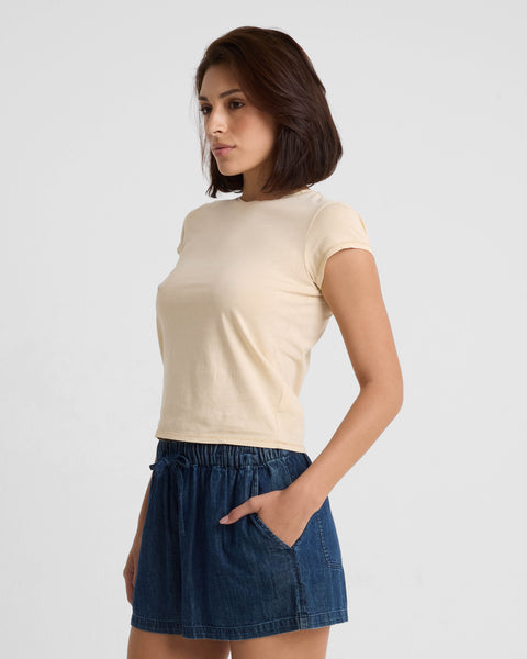A woman with short dark hair models the Volcom Women's Lived In Stone Wash Short Sleeve Tee and blue denim shorts, standing against a white background with her hands in her pockets, gazing to the side.