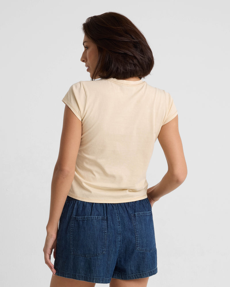 A woman with short brown hair, seen from the back, wears the Volcom Women's Lived In Stone Wash Short Sleeve Tee in beige and blue denim shorts with large back pockets against a plain white background.