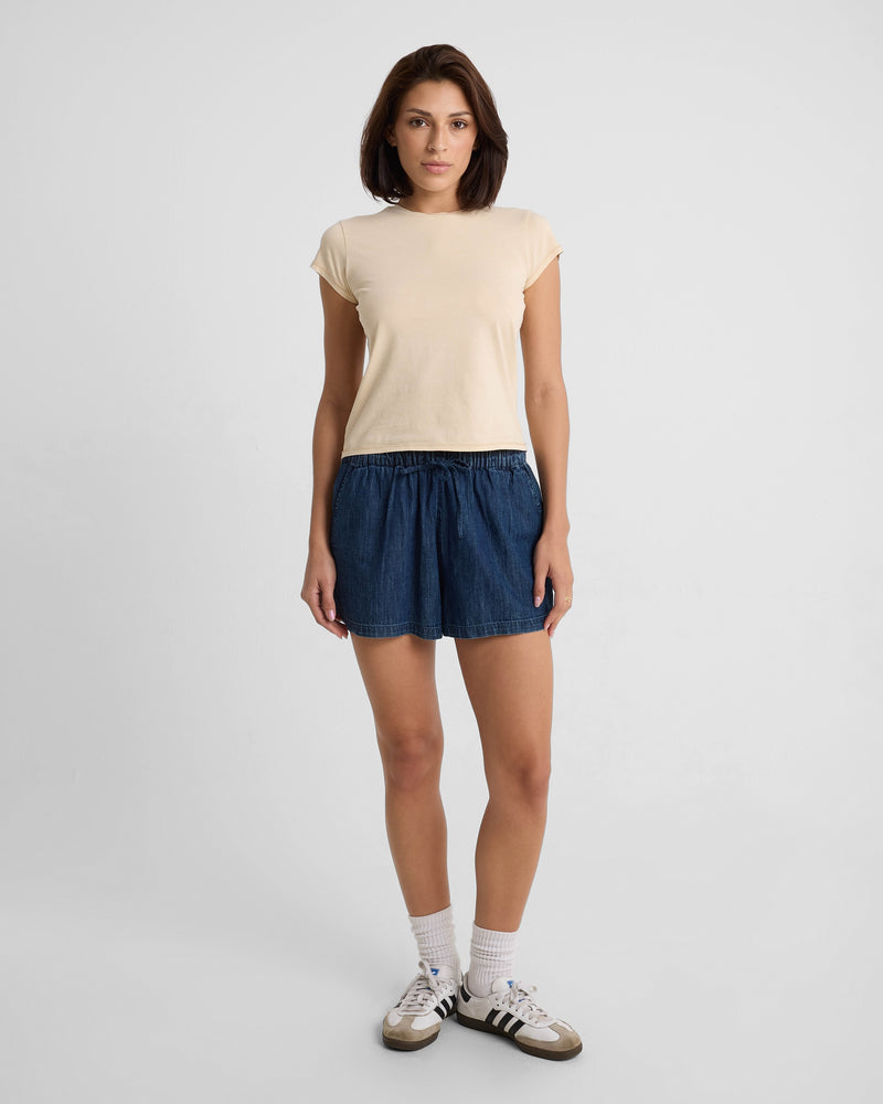 A woman faces forward in a Volcom Women's Lived In Stone Wash Short Sleeve Tee, beige color, paired with blue denim shorts, white socks, and black-and-white sneakers against a plain, light background.