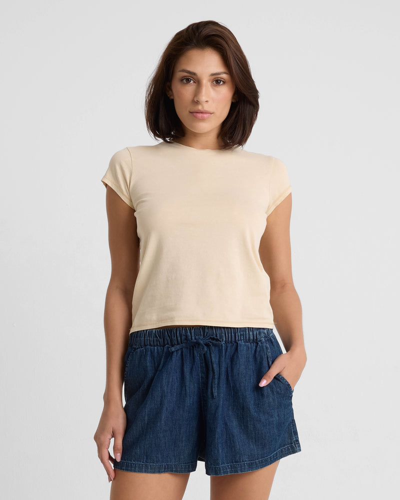 A woman with short brown hair wears the Volcom Women's Lived In Stone Wash Short Sleeve Tee and blue denim shorts with an elastic waistband, standing against a plain white background.