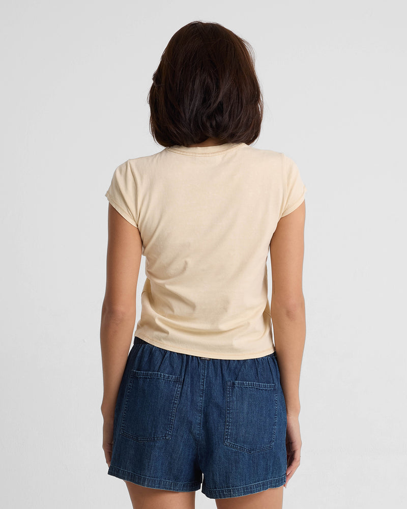A woman with short brown hair is seen from behind, wearing the Volcom Women's Lived In Stone Wash Short Sleeve Tee in light beige and blue denim shorts, standing against a plain white background.