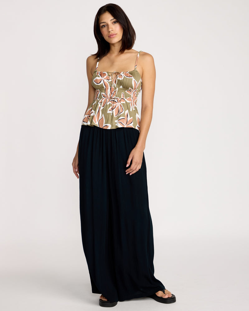 A woman faces forward in a Volcom Women's Psychonic Cami featuring adjustable straps and a smocked bodice, paired with a loose black ankle-length skirt and black sandals, against a plain white background.