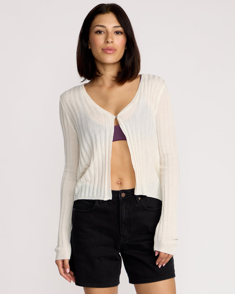 A woman with shoulder-length dark hair wears the Volcom Women's Pulp Stitchin Cardi, a white ribbed, body-skimming cardigan, layered over a purple top and black shorts against a plain light background.
